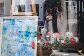 During a couple session in Hildesheim, a storefront sign reading Trust in the flow of life reflects in the window, with the couple's image softly visible behind it.